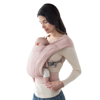 Ergobaby Embrace newborn baby carrier in blush pink, worn by a woman holding a sleeping baby.