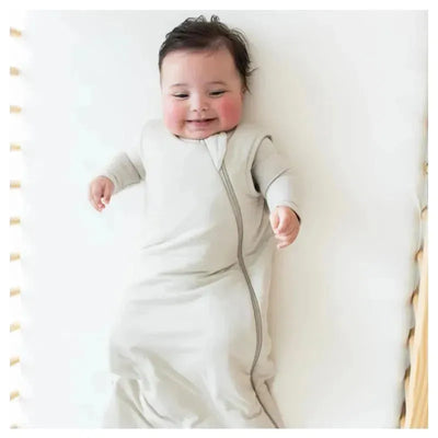 Smiling baby in a light beige Kyte Baby bamboo sleep bag, lying on a white surface with playful hands.