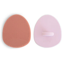 Two Mushie cradle cap brushes in rose and soft lilac colors, featuring textured surfaces for gentle cleaning.