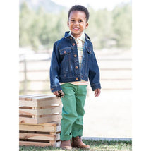 Boys denim jacket in medium wash worn by a smiling child, paired with a striped shirt, green pants, and brown boots.