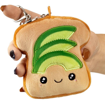 Squishable avocado toast keychain, plush with a smiling face and green avocado slices, held by a hand.