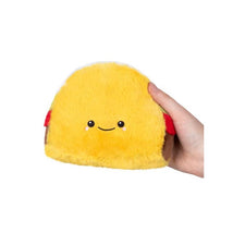 Plush taco-shaped toy with a smiling face, held in a hand, featuring yellow fur and red accents.