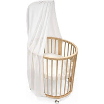 White canopy draped over a circular Stokke Sleepi crib with wooden slats and a soft mattress inside.