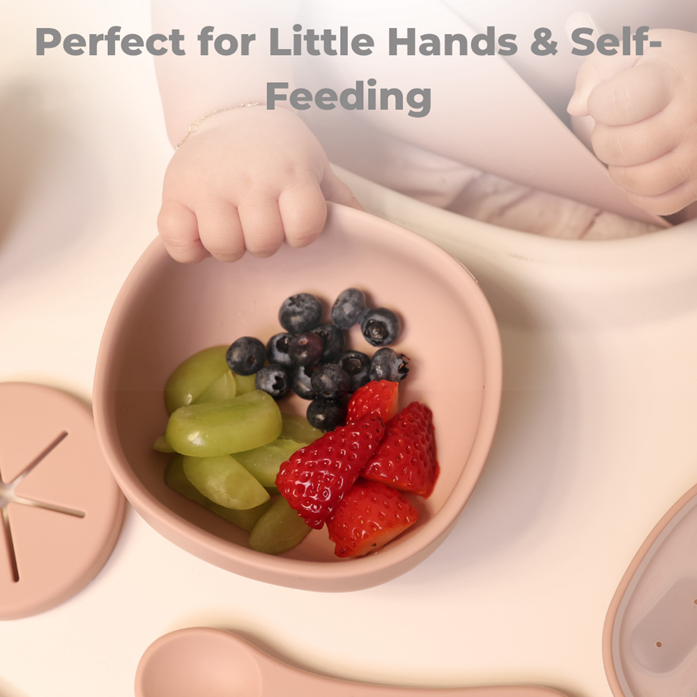 Baby self-feeding with pink silicone bowl filled with strawberries, blueberries, and grapes