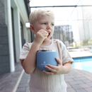 A toddler eating from a dusty blue Primo Passi insulated food jar outdoors.