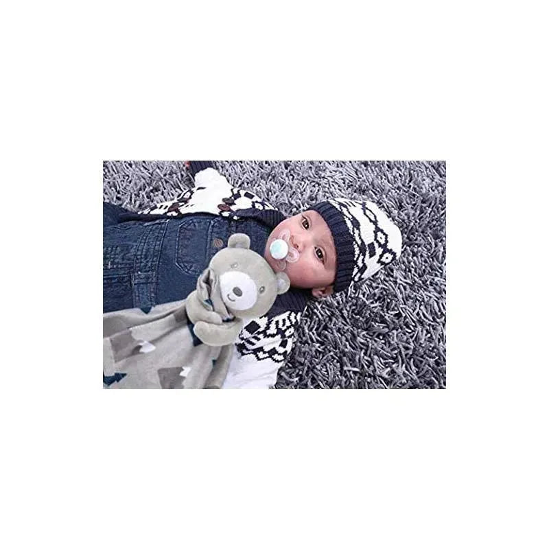 Infant wearing a knitted hat and overalls, holding a gray bear security blanket, lying on a textured rug.