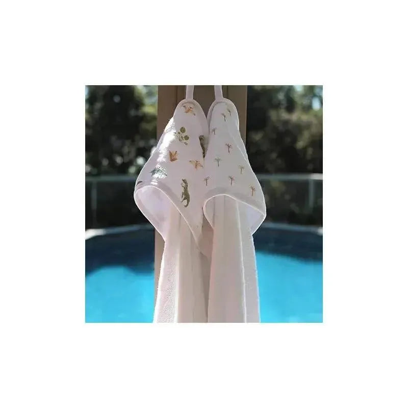 Hooded baby bath towels from Aden + Anais featuring dinosaur and jungle-themed embroidery, hung near a pool.