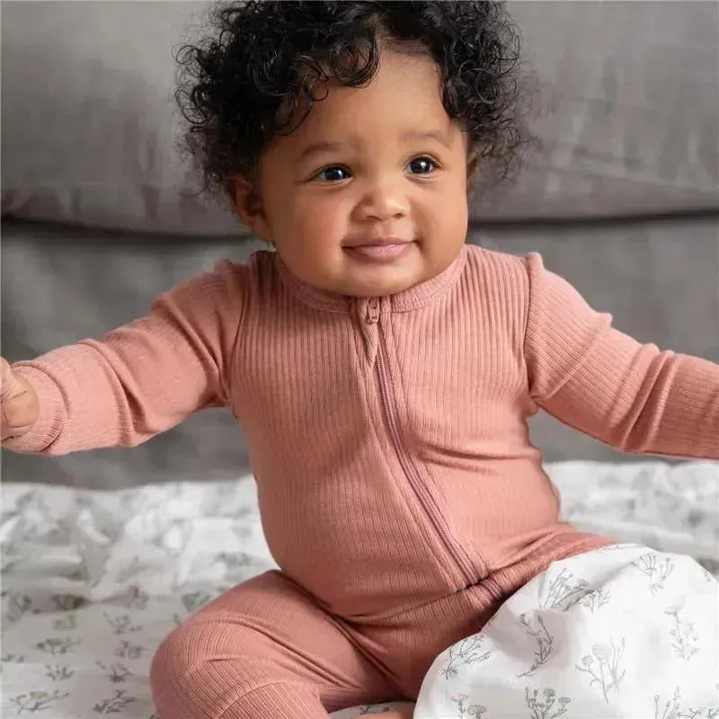 Smiling baby wearing a soft pink ribbed zip-up onesie, seated on a floral-patterned swaddle.