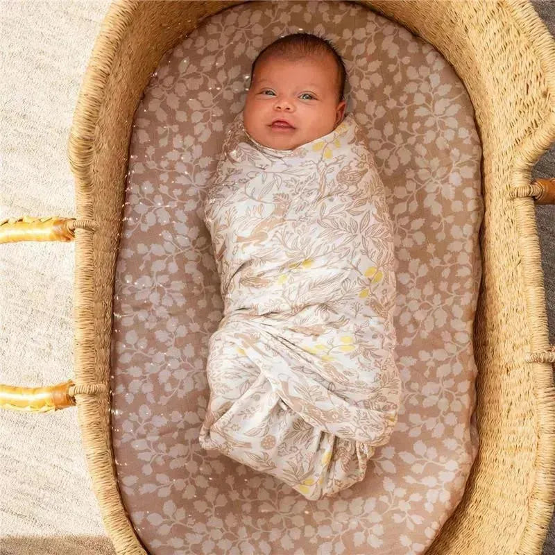 Baby swaddled in a silky soft Aden + Anais swaddle with a floral design, lying in a woven bassinet.