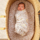 Baby swaddled in a silky soft Aden + Anais swaddle with a floral design, lying in a woven bassinet.