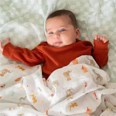 A smiling baby wearing an orange sweater, wrapped in a swaddle blanket featuring circus animals and bright colors.