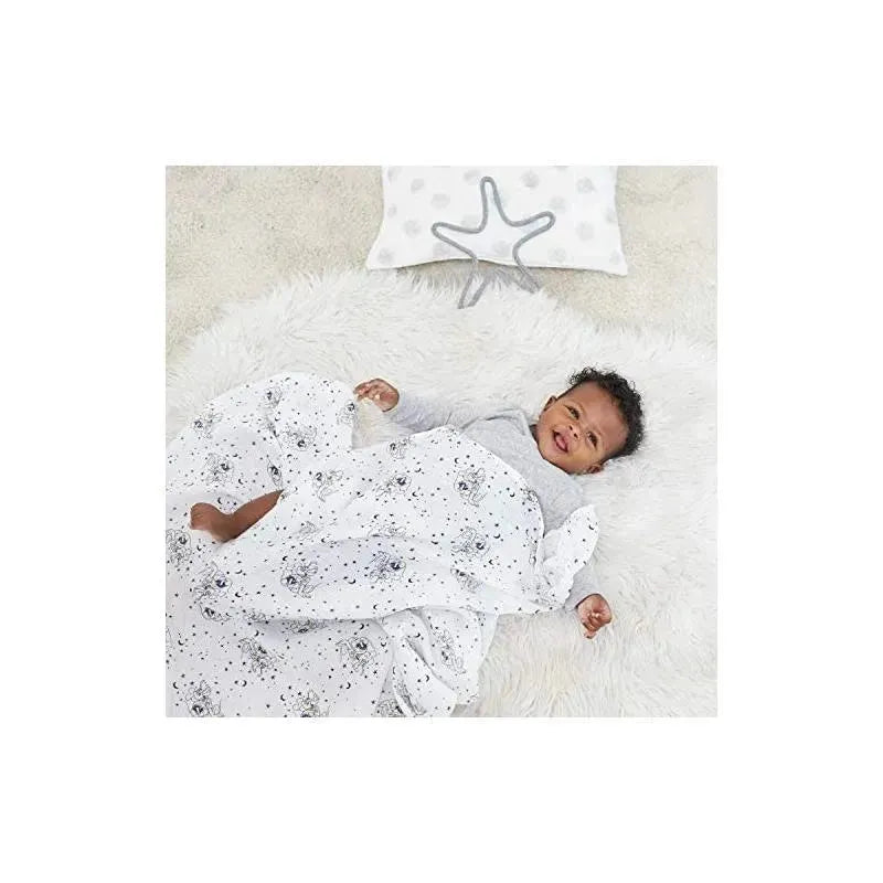 A baby lies on a soft surface, wrapped in a white muslin swaddle with a Mickey mouse stargazer print, smiling.