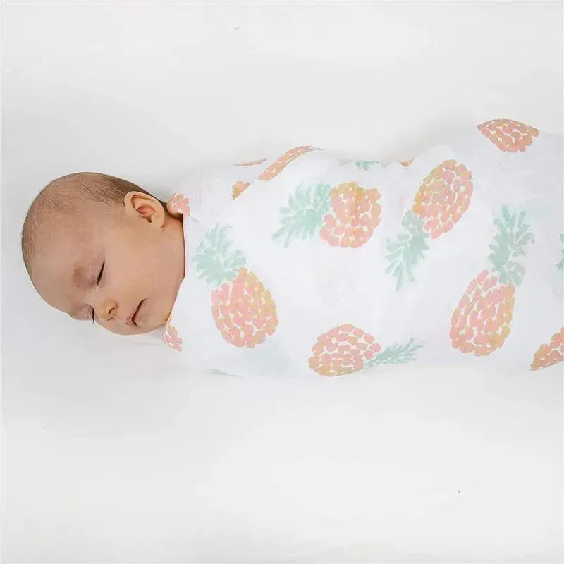 A sleeping baby is wrapped in a soft, white muslin swaddle featuring a colorful pineapple print from Aden + Anais.