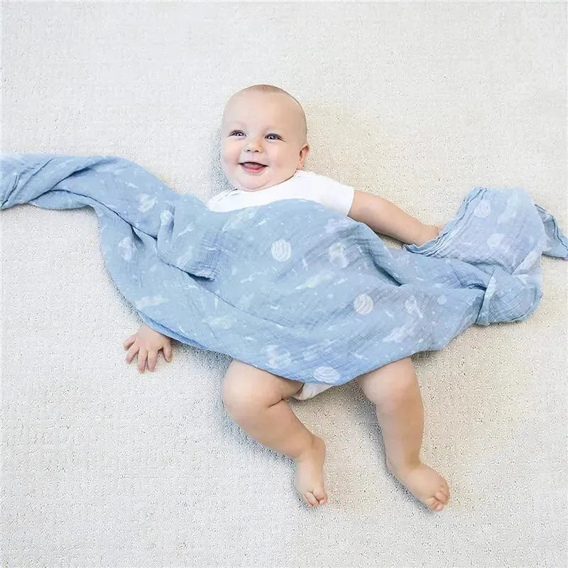 Smiling baby wearing a white onesie, playing with a blue muslin swaddle featuring space-themed patterns.