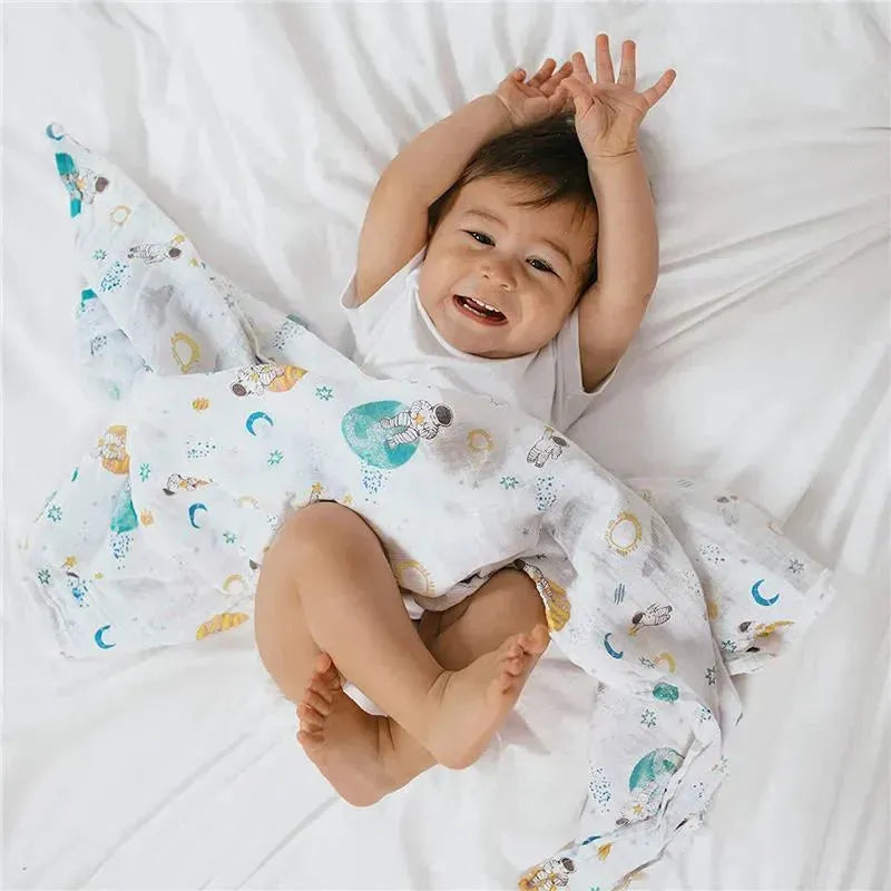 A happy baby clutches a muslin swaddle with a space explorer design, playing on a white bedspread.