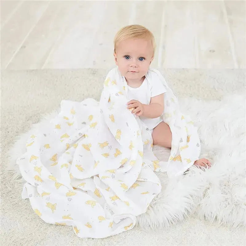 A baby sits on a rug, wrapped in a soft, white Aden+Anais blanket featuring yellow Winnie the Pooh characters.