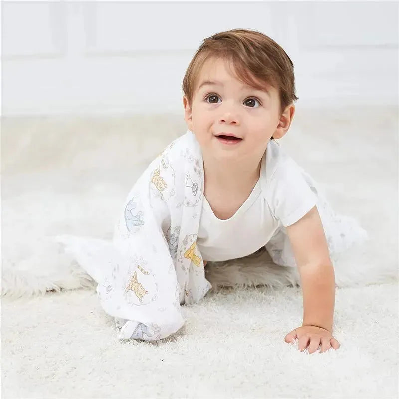 Toddler in a white onesie crawling on a soft rug, holding a lightweight Aden+Anais muslin blanket featuring Winnie the Pooh characters.