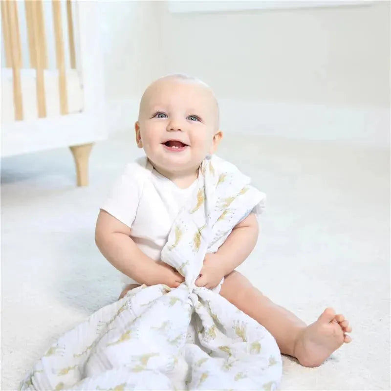 Baby sitting on the floor, smiling while holding a Aden + Anais cotton muslin swaddle featuring a natural history design.