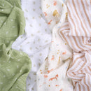Four cotton muslin swaddles in sage green, white with woodland animals, and striped patterns, showcasing soft textures.