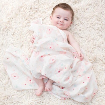 A smiling baby wrapped in a soft, floral-patterned muslin swaddle blanket lies on a fluffy rug.