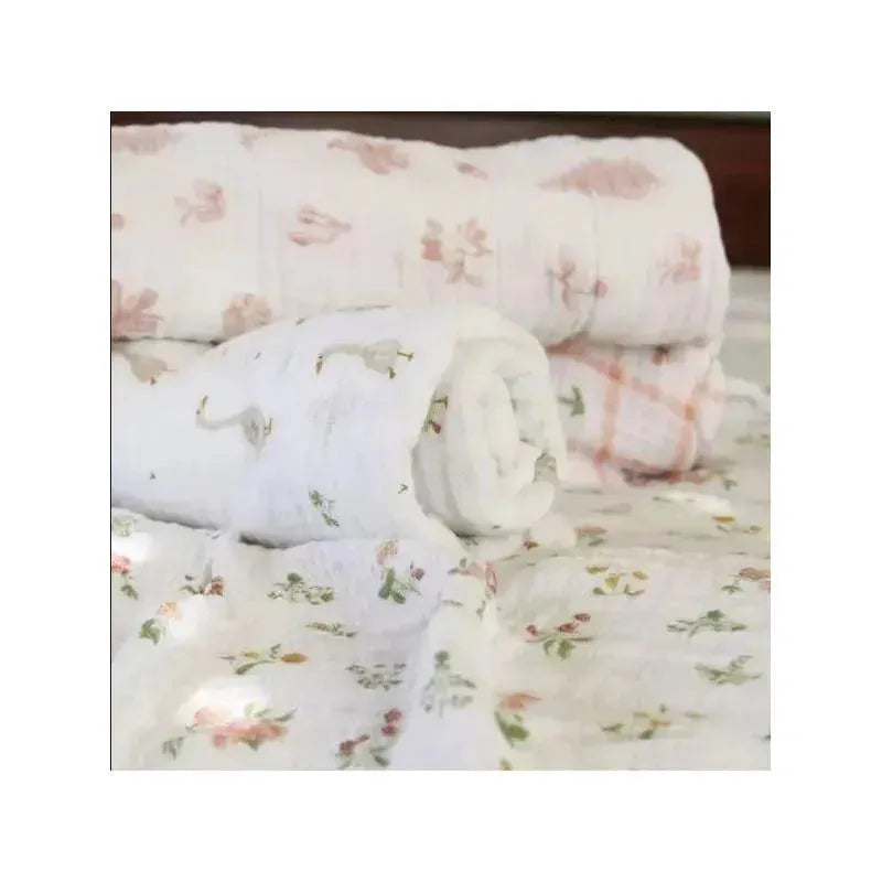 Four Aden + Anais muslin swaddle blankets in various floral and pattern designs, stacked neatly on a surface.
