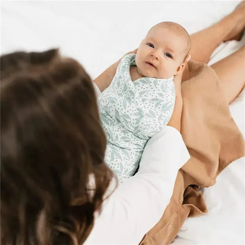 Aden + Anais muslin swaddle in leaf pattern, with a baby comfortably wrapped in it, held by a caregiver.