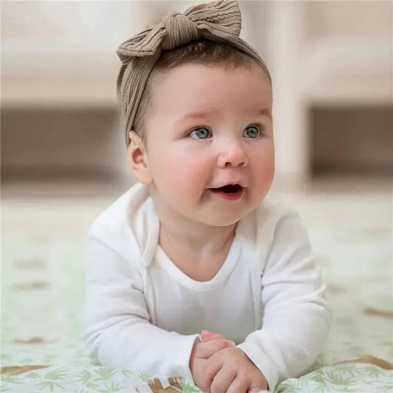 Smiling baby with light blue eyes wearing a white long-sleeve shirt and a tan knotted headband, sitting on a patterned mat.