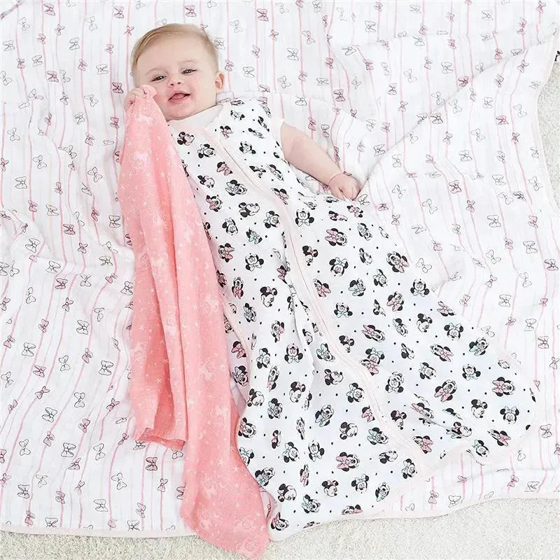 Baby lying on a muslin blanket, wearing a Minnie Mouse sleeping sack and holding a pink Aden + Anais muslin blanket.