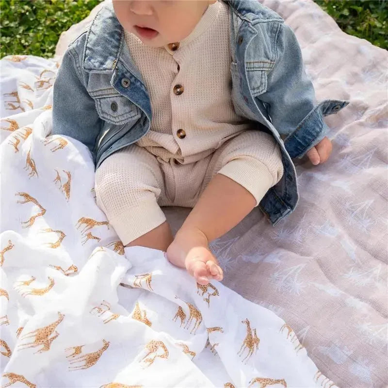 Baby wearing a denim jacket and beige outfit, sitting on a swaddle blanket featuring giraffe patterns, outdoors.