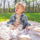 A toddler in a denim jacket sits on a light blanket surrounded by soft swaddle blankets featuring animal designs.