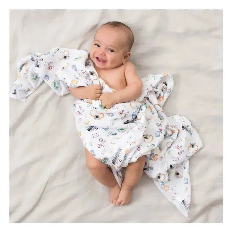 Smiling baby wrapped in a printed Aden + Anais swaddle featuring various colorful designs, lying on a light background.