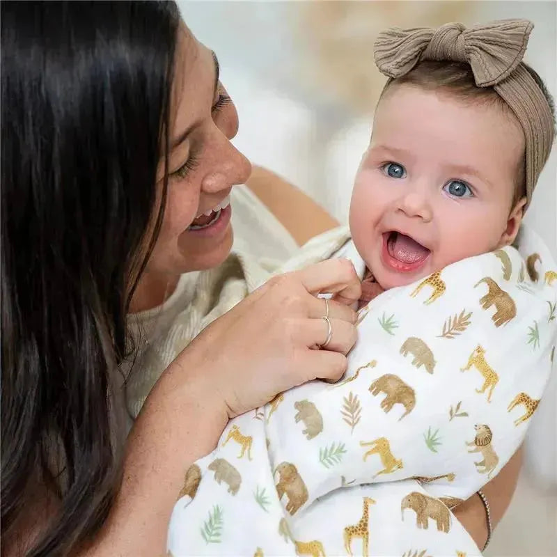 A mother smiles at her happy baby wrapped in a Aden + Anais Tanzania-Great Migration Blanket featuring safari animals.