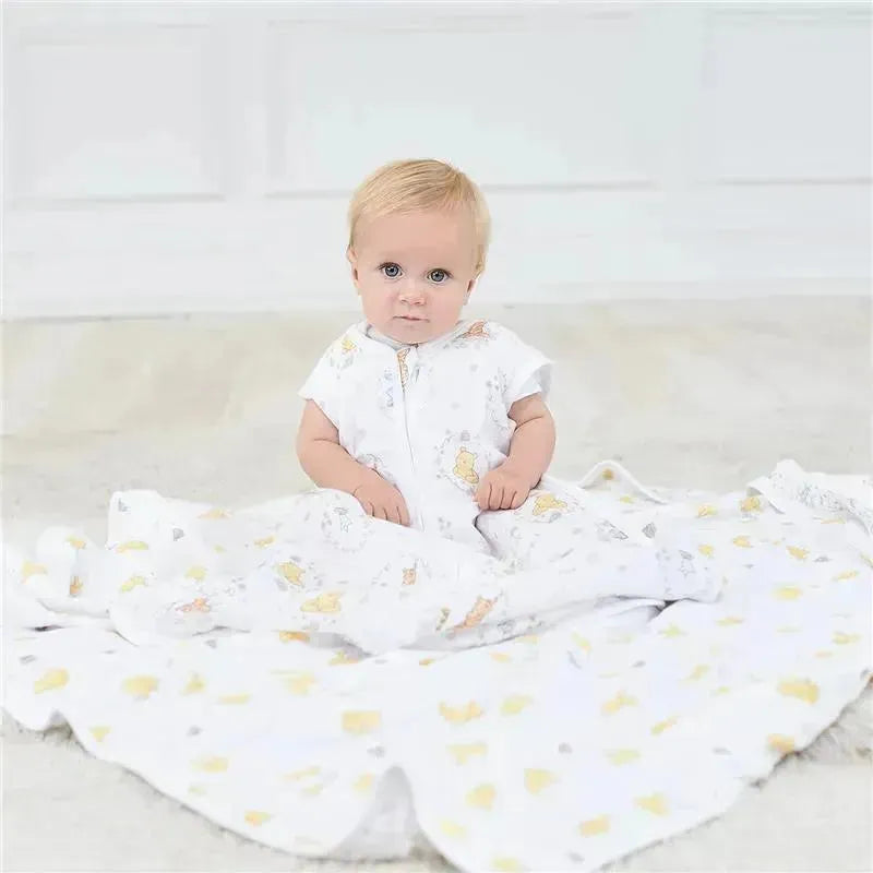 Toddler sits on a soft surface wearing a white Aden + Anais sleeping bag with a yellow and gray animal pattern.