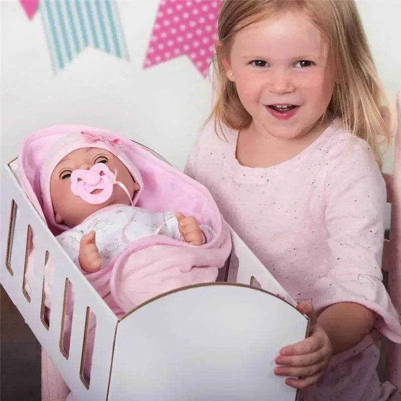 A girl smiles while holding a white crib with a baby doll wrapped in pink, featuring a pacifier and sleepwear.