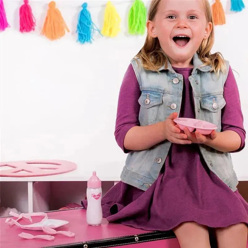 Smiling girl holding a pink plate, surrounded by a pink bottle and a colorful tassel backdrop, showcasing the Adora Magic Feeding Set.