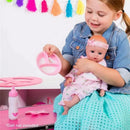 A girl in a denim jacket holds a baby doll and a pink feeding set, including a bottle, in a colorful playroom.