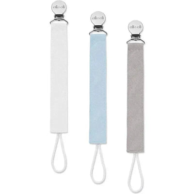 Three pacifier clips from Ali + Oli in linen material: one white, one sky blue, and one gray, each with a metal clasp.