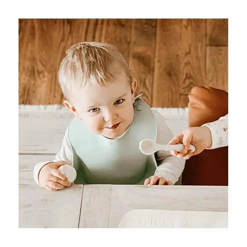 Light sage silicone baby bib on a smiling toddler at a wooden table, with a hand offering a spoon.