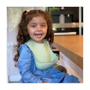 Smiling young girl with curly hair wearing a light sage silicone baby bib, seated at a kitchen counter.