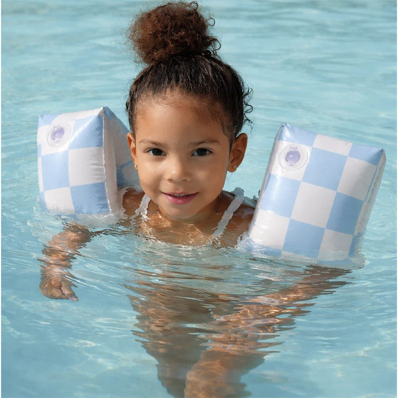 Child smiling and floating in water, wearing blue checkered inflatable swim arm bands.