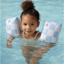 Child smiling and floating in water, wearing blue checkered inflatable swim arm bands.
