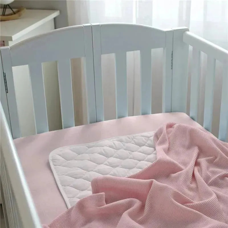 Fitted mini crib sheet in baby pink placed on a crib mattress, with a white quilted mattress protector and soft blanket.