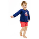 Baby wearing a navy sweatshirt with a sailboat applique and red paperbag shorts, smiling and posing.