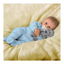 Baby boy doll dressed in a blue knitted outfit, holding a plush dog and a pacifier, resting on a soft blanket.