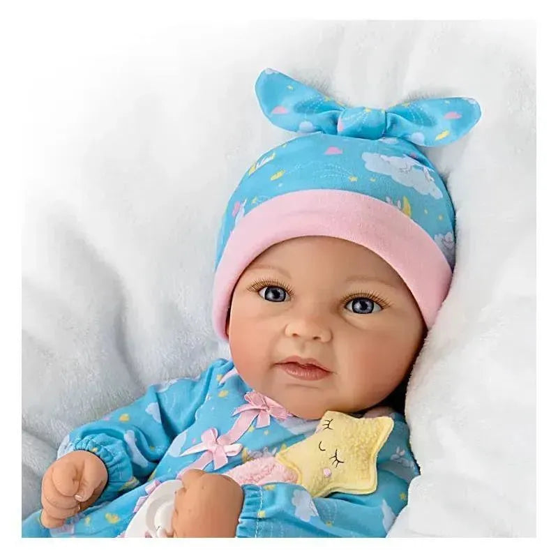 Ashton Drake Baby Doll Rylee, wearing a blue pajama and pink hat, holding a star-shaped toy, is nestled in a soft white blanket.