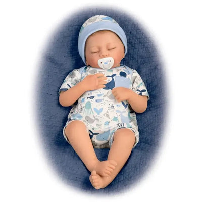 Breathing baby boy doll wearing a blue hat and patterned outfit, nestled on a quilted blanket with a pacifier.