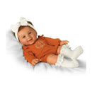 Lilah Baby Girl doll wearing an orange romper with white ruffled socks and a bow headband, smiling on a soft surface.