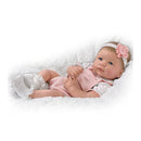 Lifelike silicone baby doll named Ava by Ashton Drake, wearing a pink outfit with a floral headband, resting on soft white bedding.
