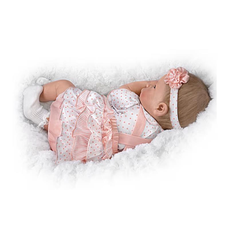 Lifelike silicone baby doll by Ashton Drake, featuring a polka dot dress, pink accents, and a floral headband, resting on fluffy white fabric.