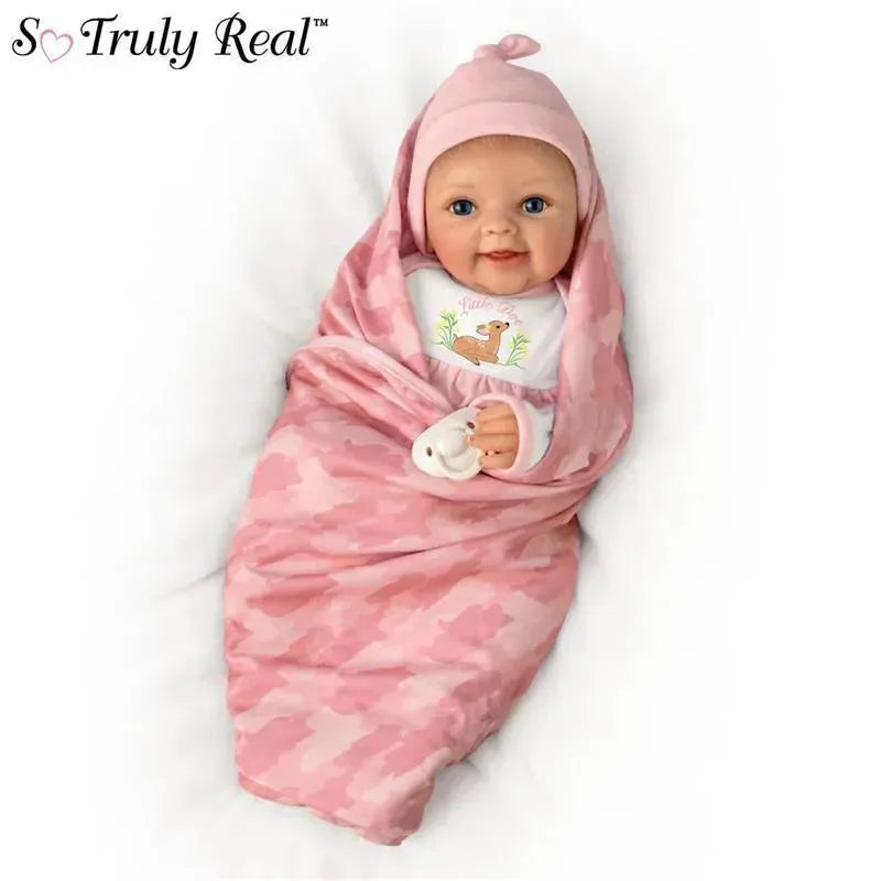 Lifelike baby girl doll wrapped in a pink camouflage blanket, wearing a matching hat and a shirt featuring a fawn illustration.